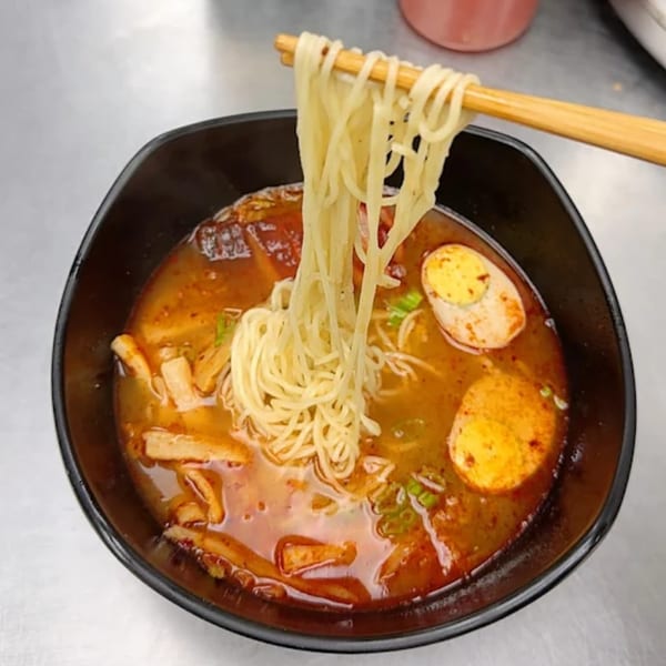 Chopsticks lifting noodles from a bowl of spicy Asian ramen with eggs and meat.