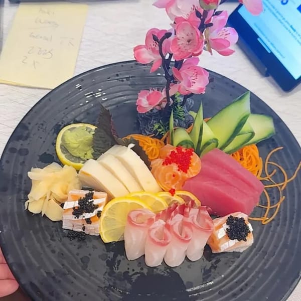 A black plate of Asian Arch sashimi and sushi with lemon, ginger, wasabi, and pink floral decor.