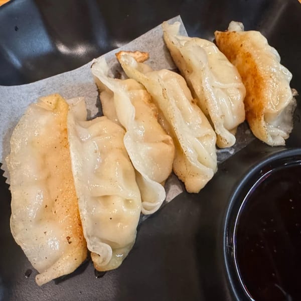 Six pan-fried Asian dumplings with crispy bottoms and a side of dark dipping sauce.